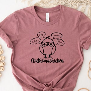 May include: A mauve t-shirt with a black graphic of a cartoon chicken wearing glasses and thought bubbles with math equations. The text "Mathemachicken" is below the chicken.