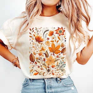 May include: Cream-colored t-shirt featuring a rectangular floral design. The design showcases autumn-toned flowers and leaves in shades of orange, brown, and yellow. The shirt is paired with blue jeans.