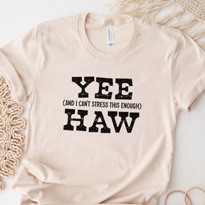 Op de afbeelding: Een beige T-shirt met de tekst "YEE (AND I CAN'T STRESS THIS ENOUGH) HAW" in zwart.