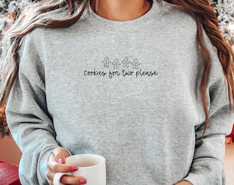 May include: A heather gray crewneck sweatshirt featuring an embroidered design of four small gingerbread men above the dark green text that reads, "cookies for two please." The garment has long sleeves and a ribbed neckline, cuffs, and hem.