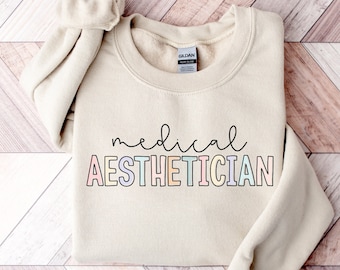 Może przedstawiać: Beżowa bluza z napisem "medical AESTHETICIAN" w kolorach tęczy.