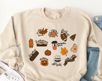 May include: A beige sweatshirt with a fall-themed design featuring illustrations of leaves, a pumpkin, a football, a coffee cup, a scarf, and the words "falling leaves", "sweatshirt", "football", "pumpkin spice", "coffee dates", "cooler weather", "bonfire", "Thanksgiving", "fall"
