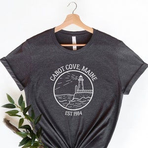 Puede incluir: Camiseta gris oscuro con un gr&aacute;fico blanco de un faro en un c&iacute;rculo con el texto "Cabot Cove, Maine Est 1984".