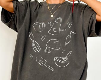 May include: A dark gray t-shirt with a white line drawing of kitchen tools and hearts. The design includes a whisk, mixer, spatula, bowl, and other baking-related items. The shirt has short sleeves and a relaxed fit.