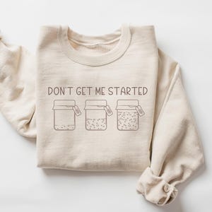 Peut inclure: Un sweat-shirt beige avec le texte "Don't get me started" et un graphique de trois bocaux avec des couvercles. Chaque bocal a une &eacute;tiquette qui dit "Good Vibes Only".
