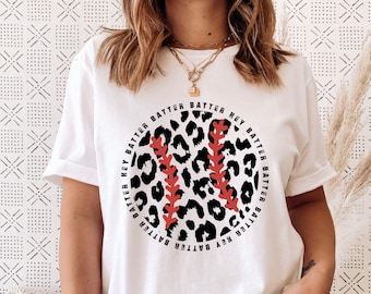 May include: White t-shirt with a black and white leopard print baseball graphic. The graphic has red stitching and the words "Batter Up" repeated around the outside of the baseball.