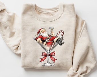 May include: A beige crewneck sweatshirt featuring a graphic illustration of Santa Claus relaxing inside a large martini glass. Santa wears a red suit and black boots, holding a martini glass with an olive, while a candy cane rests nearby.