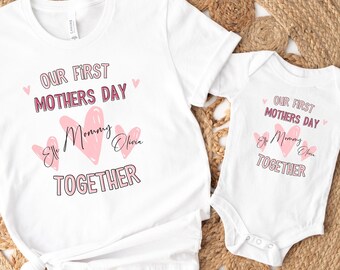 May include: White t-shirt and onesie set with the text "OUR FIRST MOTHERS DAY" and "Ellie Mommy Olivia TOGETHER" in pink. The design includes pink hearts. The set is displayed on a woven, circular mat.