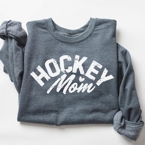 May include: A dark gray sweatshirt with the text "HOCKEY MOM" in white with a distressed look. The sweatshirt is folded and laid flat on a white surface.