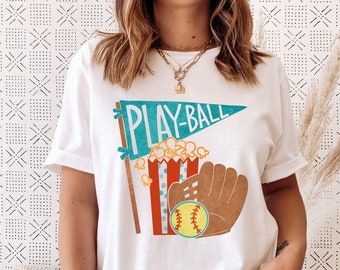 May include: White t-shirt with a graphic design of a blue pennant with the text "PLAY-BALL", a red and white striped popcorn bucket with popcorn, and a brown baseball glove with a yellow baseball.