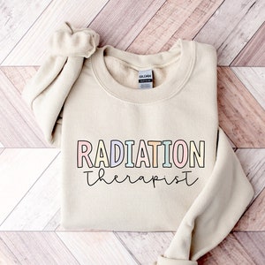 Pu&ograve; includere: Una felpa beige con il testo "RADIATION therapist" in colori arcobaleno. Il testo &egrave; contornato di nero.