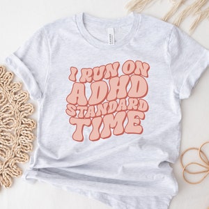 May include: A grey t-shirt with the text "I run on ADHD standard time" in pink letters. The letters are in a retro font style.