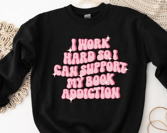 May include: A black long-sleeve crewneck sweatshirt featuring the pink text I WORK HARD SO I CAN SUPPORT MY BOOK ADDICTION in a bold, rounded font, accented with small white star graphics surrounding the lettering.