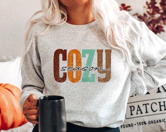 May include: A gray sweatshirt with the text "Cozy season" in colorful retro-style lettering. The sweatshirt is being worn by a person holding a black mug.