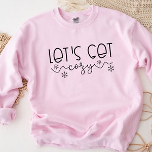 May include: A pink sweatshirt with the text "Let's Get Cozy" in black lettering with snowflake accents.