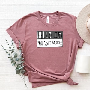 May include: A dusty rose t-shirt with a name tag design that says "Hello I'm Probably Anxious".