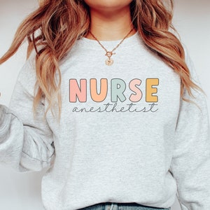 May include: A light gray sweatshirt with the text "NURSE anesthetist" in colorful letters. The letters are outlined in black and have a rounded, playful font.
