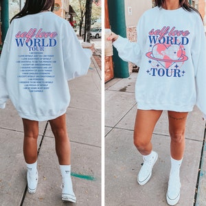 May include: White sweatshirt with a blue and pink graphic print that says "self love world tour" and includes a heart with a globe inside. The text on the back of the sweatshirt says "self love world tour" and includes a list of positive affirmations.