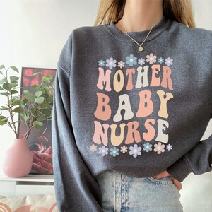 May include: A dark gray sweatshirt with the text "MOTHER BABY NURSE" in colorful retro-style lettering. The text is surrounded by a row of small pastel flowers.