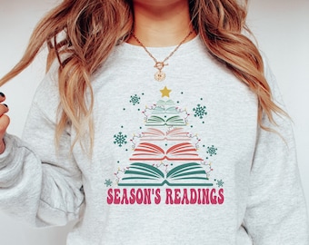 Könnte beinhalten: Ein graues Sweatshirt mit einem Weihnachtsbaumdesign aus gestapelten Büchern. Der Baum ist mit Lichtern und Schneeflocken verziert. Der Text "Season's Readings" ist unter dem Baum gedruckt.