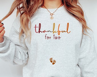 Peut inclure: Un sweat-shirt gris avec le texte "thankful for two" en dégradé arc-en-ciel. Le texte est en écriture cursive et il y a deux petites empreintes de pas brunes sous le texte.
