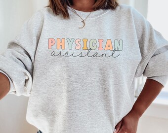 May include: A light gray sweatshirt with the words "PHYSICIAN ASSISTANT" printed across the front in colorful block letters and a cursive font. The sweatshirt has long sleeves and a crew neck.