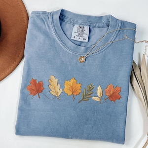 May include: A light blue t-shirt with a design of five autumn leaves in shades of red, yellow, orange, and brown. The leaves are connected by a thin line. The shirt has a tag that reads "Comfort Colors" and "S - P - CH".