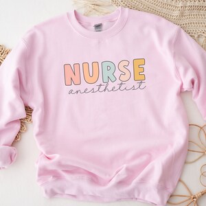 May include: Pink sweatshirt with the text "NURSE anesthetist" in colorful letters.