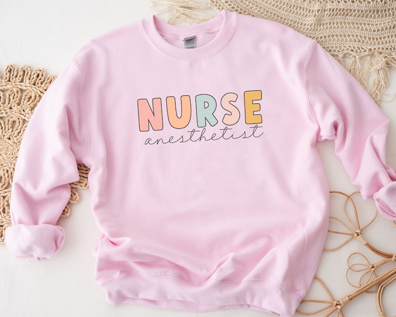 May include: Pink sweatshirt with the text "NURSE anesthetist" in colorful letters.