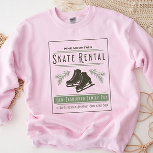 K&ouml;nnte beinhalten: Rosafarbener Pullover mit einem grafischen Design eines Vintage-Schilds mit der Aufschrift "Pine Mountain Skate Rental" mit einem Paar Schlittschuhen und Kiefern&auml;sten. Der Text unter dem Schild lautet "Old-Fashioned Family Fun" und "20 All-Day Rentals - Open Daily - Cocoa & Hot Cider".