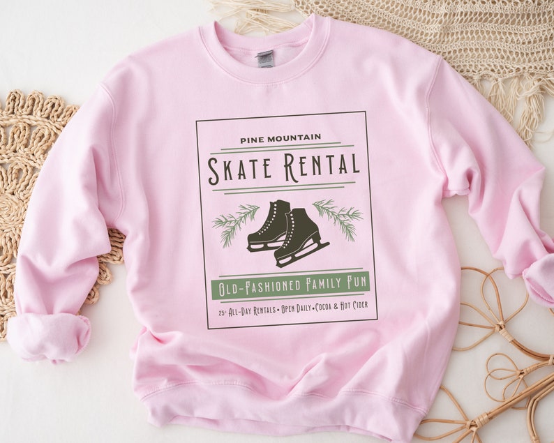K&ouml;nnte beinhalten: Rosafarbener Pullover mit einem grafischen Design eines Vintage-Schilds mit der Aufschrift "Pine Mountain Skate Rental" mit einem Paar Schlittschuhen und Kiefern&auml;sten. Der Text unter dem Schild lautet "Old-Fashioned Family Fun" und "20 All-Day Rentals - Open Daily - Cocoa & Hot Cider".