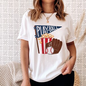 May include: White t-shirt with a blue and red pennant that says "Play-Ball" and a graphic of a popcorn bucket, baseball glove, and baseball.