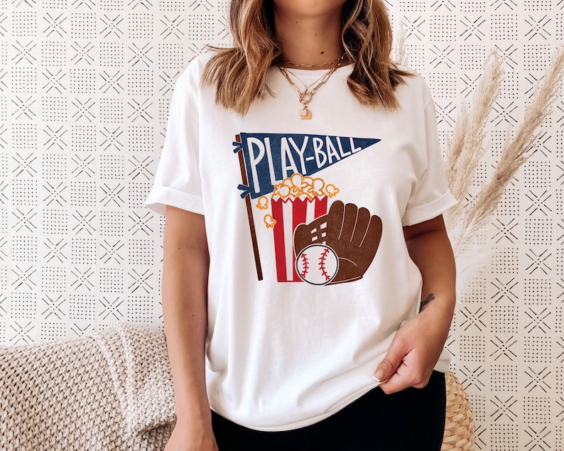 May include: White t-shirt with a blue and red pennant that says "Play-Ball" and a graphic of a popcorn bucket, baseball glove, and baseball.