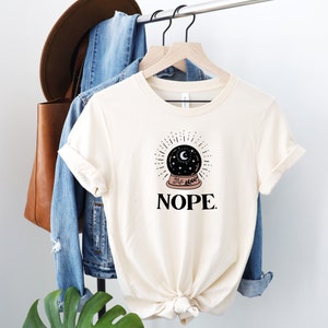 May include: A white t-shirt with a black and white illustration of a crystal ball with a crescent moon and stars inside. The text "NOPE." is printed below the illustration.