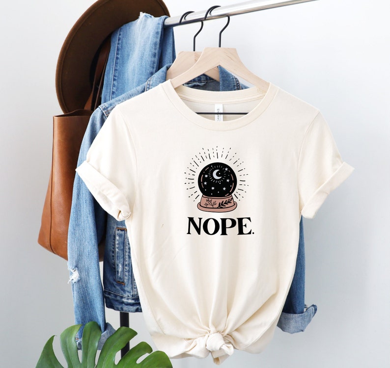 May include: A white t-shirt with a black and white illustration of a crystal ball with a crescent moon and stars inside. The text "NOPE." is printed below the illustration.