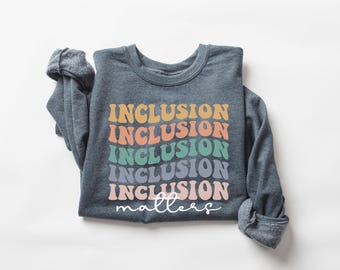 May include: A folded, long-sleeved, dark gray sweatshirt with the word "INCLUSION" printed in a retro font in multiple colors. The word "matters" is written in cursive below the word "INCLUSION".