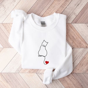 Peut inclure: Sweat-shirt blanc avec un dessin au trait noir d'un chat avec un c&oelig;ur rouge sur la queue.
