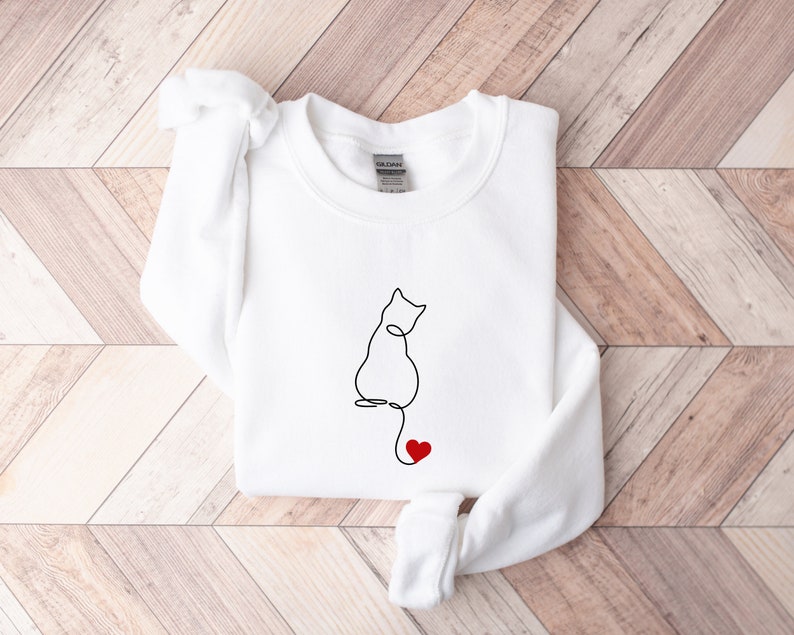 Peut inclure: Sweat-shirt blanc avec un dessin au trait noir d'un chat avec un c&oelig;ur rouge sur la queue.