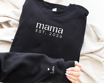 May include: A black crewneck sweatshirt featuring the white text mama EST. 2026 printed on the chest. The left sleeve cuff displays a small white heart icon followed by the name Liam in a script font, positioned near the wrist area.