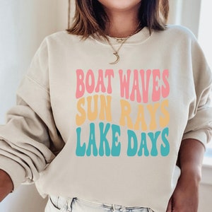 May include: A light beige sweatshirt with the text "Boat Waves Sun Rays Lake Days" in a retro font with pink, yellow, and blue colors.