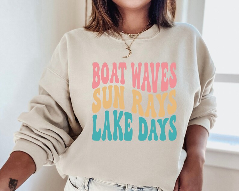 May include: A light beige sweatshirt with the text "Boat Waves Sun Rays Lake Days" in a retro font with pink, yellow, and blue colors.