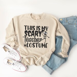 May include: A beige sweatshirt with the text "THIS IS MY SCARY Teacher COSTUME" in black, featuring a skeleton graphic. The sweatshirt is paired with blue jeans and white sneakers.