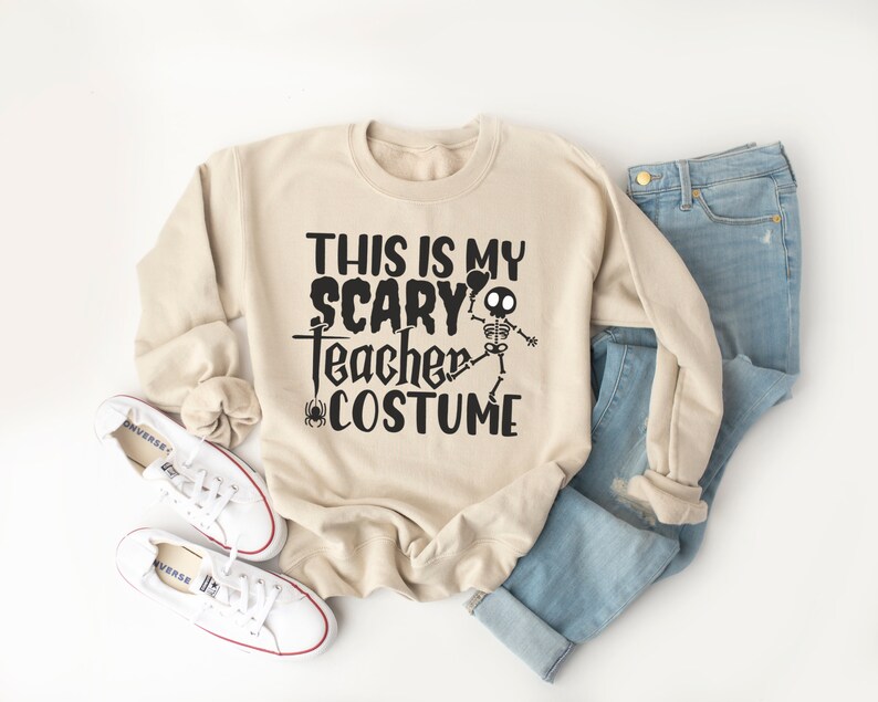 May include: A beige sweatshirt with the text "THIS IS MY SCARY Teacher COSTUME" in black, featuring a skeleton graphic. The sweatshirt is paired with blue jeans and white sneakers.