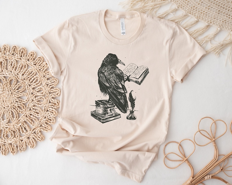Puede incluir: Una camiseta beige con una ilustraci&oacute;n en blanco y negro de un cuervo posado sobre un libro, sosteniendo una pluma en su pico. El cuervo est&aacute; leyendo un libro con el texto "The Raven" visible.