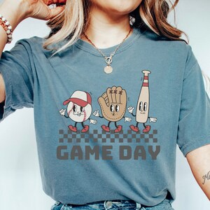 May include: A blue t-shirt featuring a graphic of a baseball, a baseball glove, and a baseball bat with cartoon faces. The graphic is on a chequered background with the text "GAME DAY" below it.