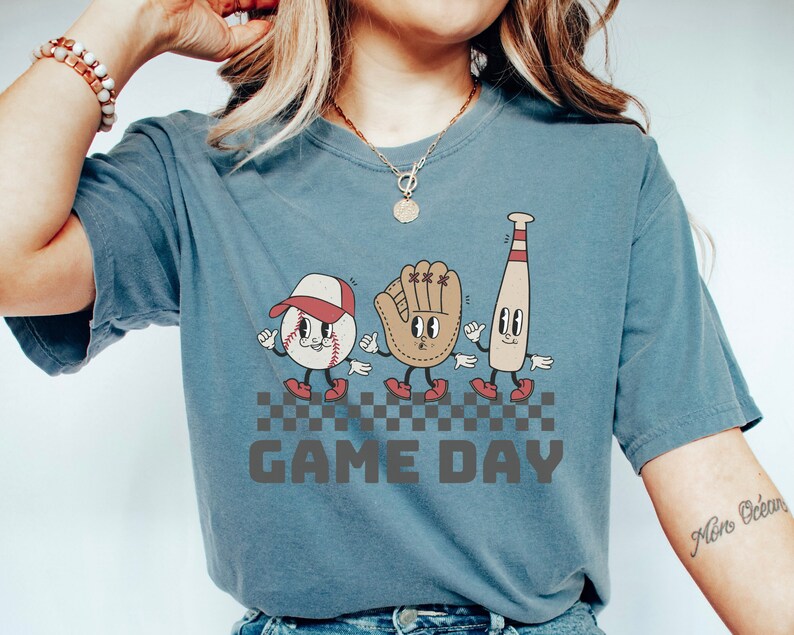 May include: A blue t-shirt featuring a graphic of a baseball, a baseball glove, and a baseball bat with cartoon faces. The graphic is on a chequered background with the text "GAME DAY" below it.
