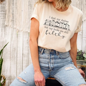 Peut inclure: Un t-shirt beige avec un motif noir qui dit "I dust my shelves but my books are still filthy". Le motif repr&eacute;sente une &eacute;tag&egrave;re avec des livres dessus.