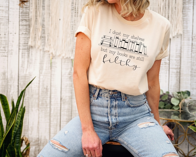 Peut inclure: Un t-shirt beige avec un motif noir qui dit "I dust my shelves but my books are still filthy". Le motif repr&eacute;sente une &eacute;tag&egrave;re avec des livres dessus.