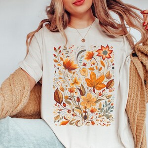 May include: A white t-shirt with a floral print featuring orange, yellow, and green flowers. The flowers are arranged in a bouquet-like design.