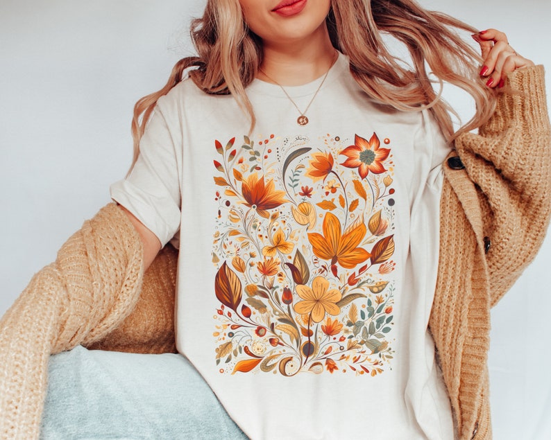May include: A white t-shirt with a floral print featuring orange, yellow, and green flowers. The flowers are arranged in a bouquet-like design.